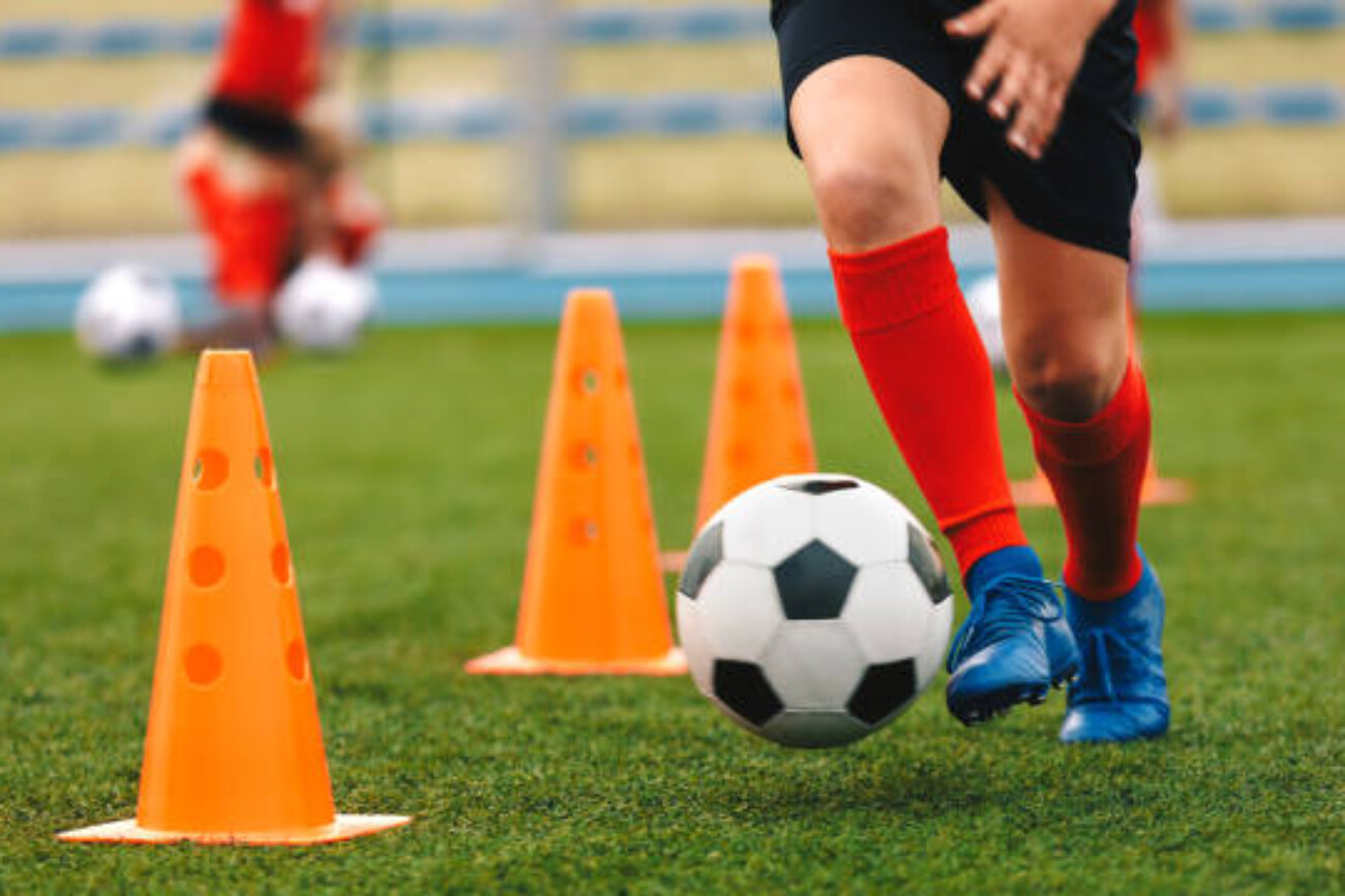 Footballer dribbling ball on training between orange cones. Young football player in sports blue cleats and red socks